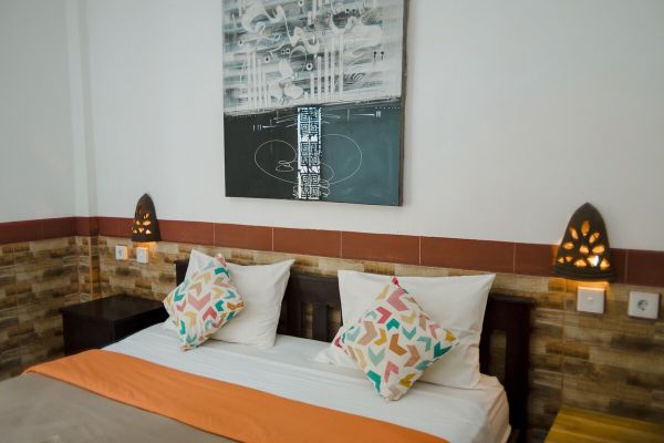 Cozy tropical bedroom with a wooden double bed, orange sheets, patterned pillows, abstract black-and-white wall art, and ornate wall sconces. - Bali Villas